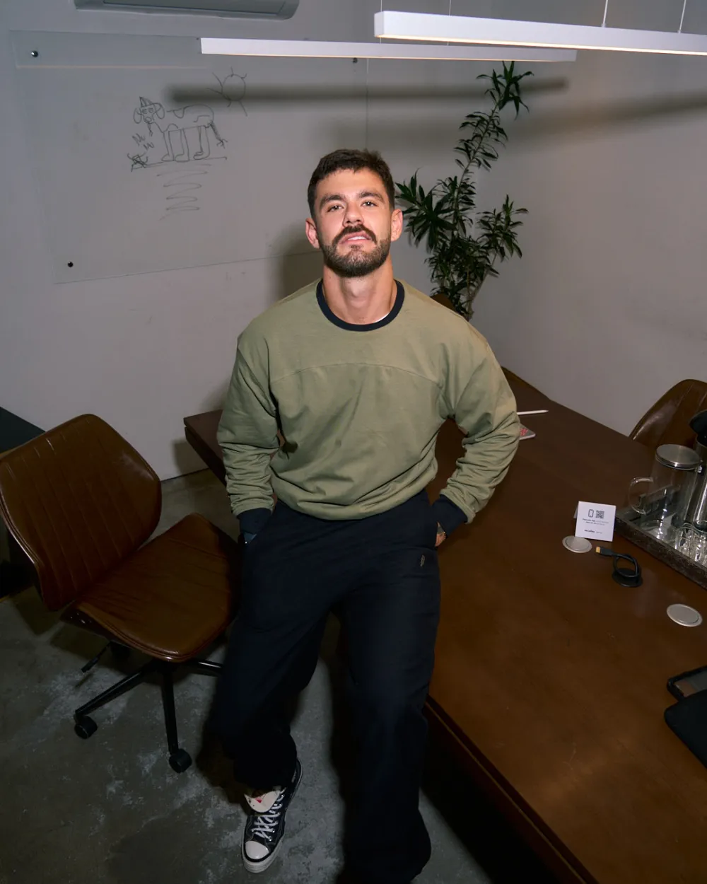 Homem de pé numa sala de reunião, de frente, com o moletom verde oliva e as mãos nos bolsos.