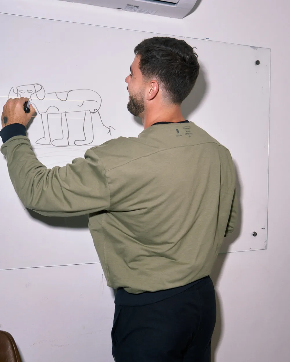 Homem escrevendo num quadro branco de costas, vestindo o moletom verde oliva.
