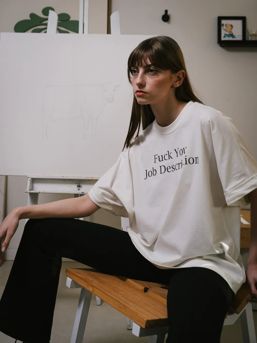 Pessoa sentada em um banco de ateliê, vestindo a camiseta, com um cavalete e o desenho de uma vaca ao fundo.