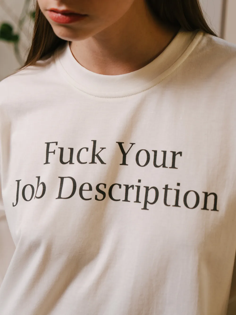 Close de uma camiseta off-white com “Fuck Your Job Description” estampado em serifa no peito.