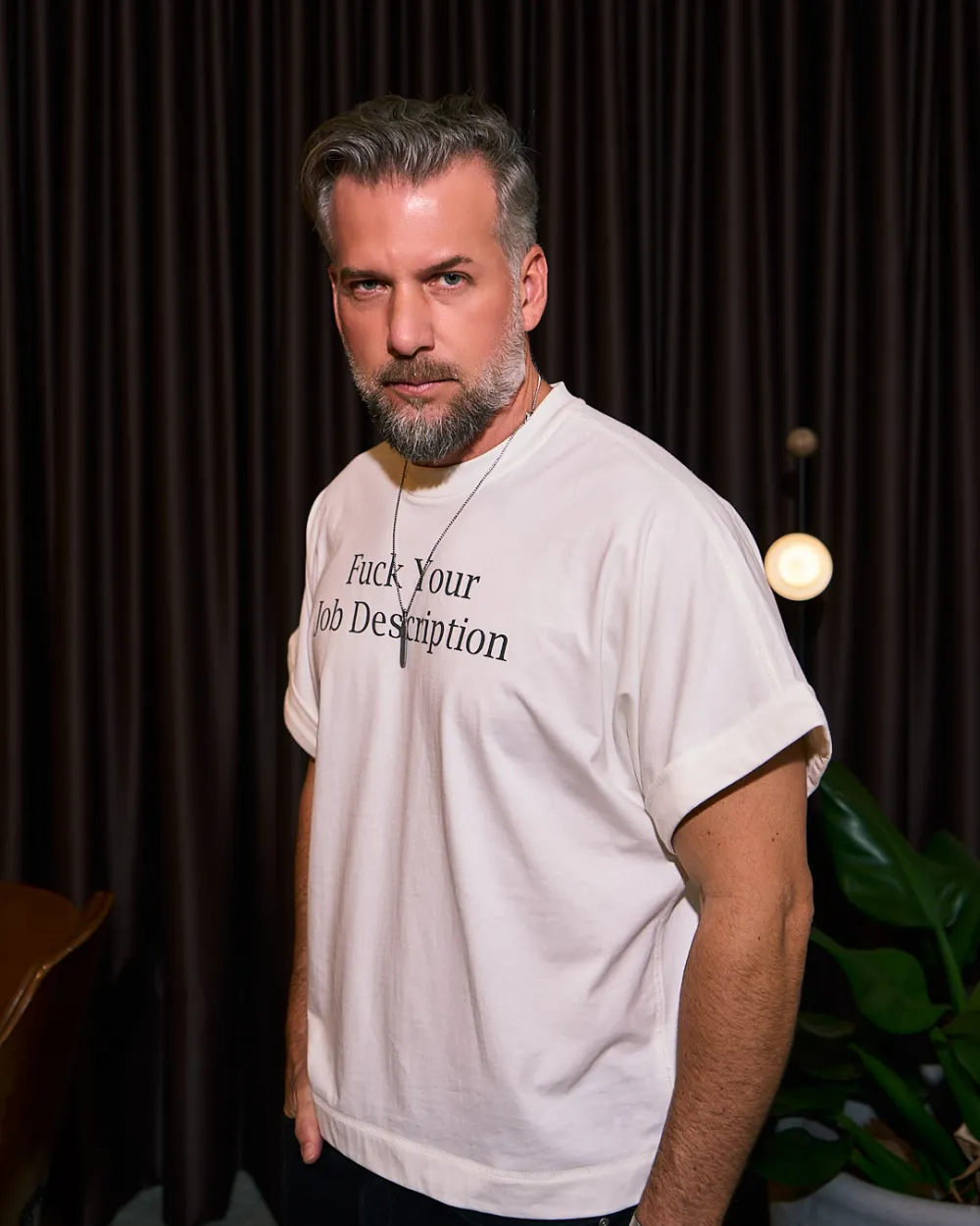 Homem de barba grisalha vestindo a mesma camiseta, de frente, contra um fundo escuro.
