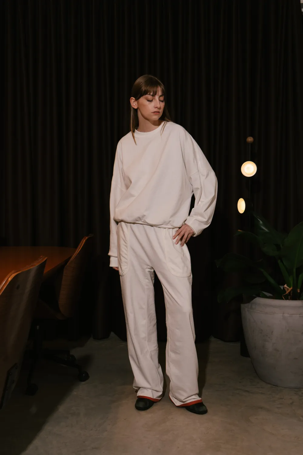 Corpo inteiro com o conjunto off-white, de pé, contra um fundo escuro.