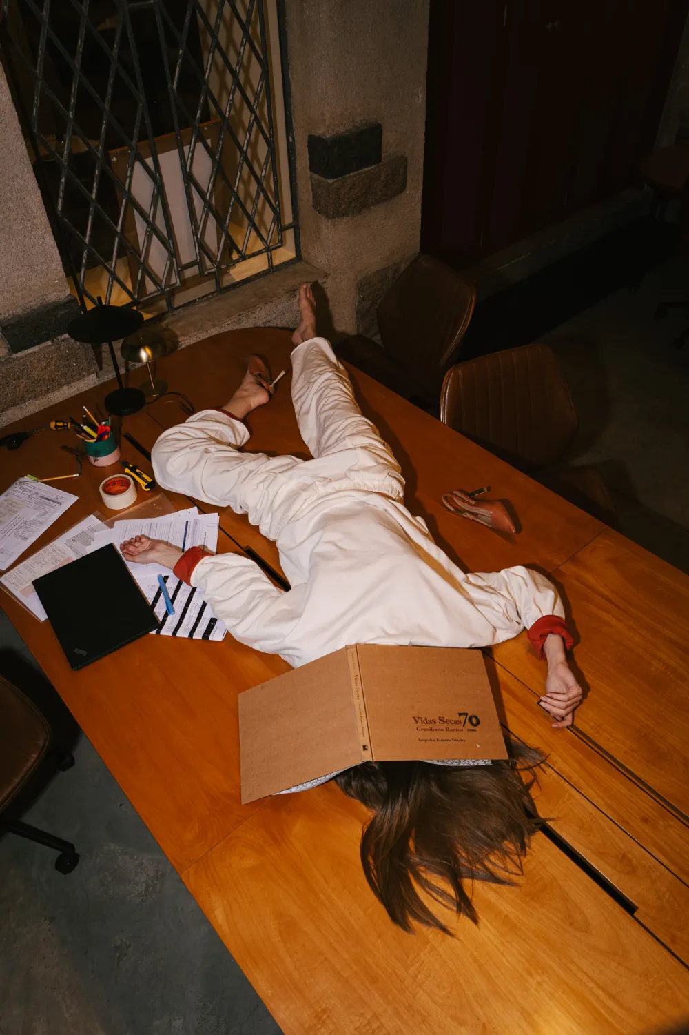 Pessoa deitada sobre uma mesa de reunião com o conjunto off-white, o braço estendido sobre os papéis.