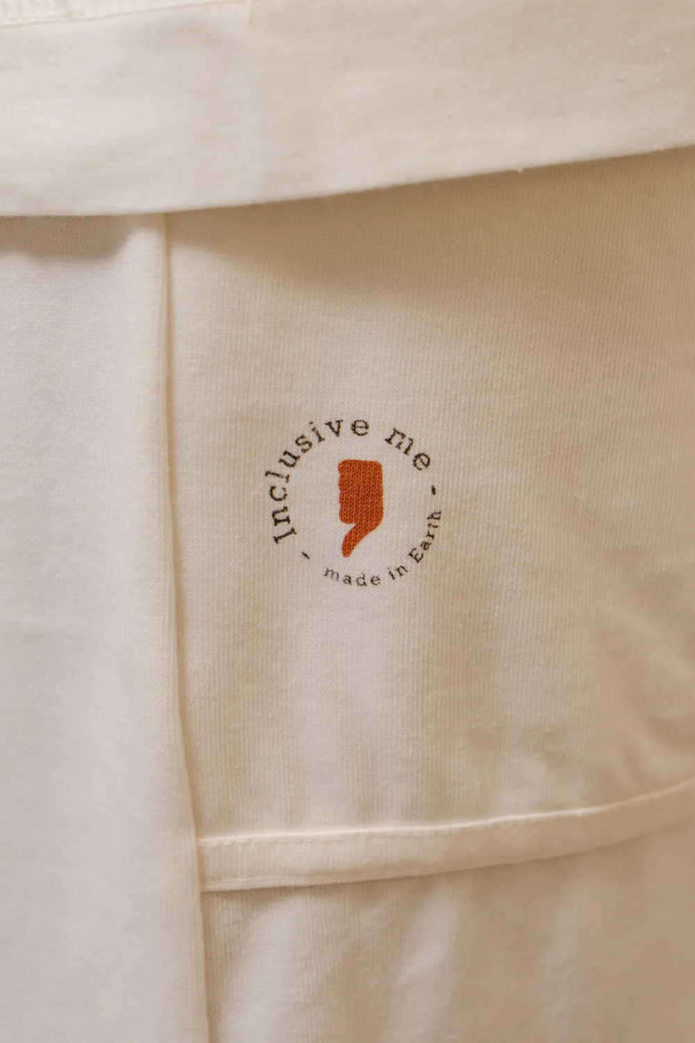 Selo “inclusive me · made in earth” estampado na barra da peça off-white.