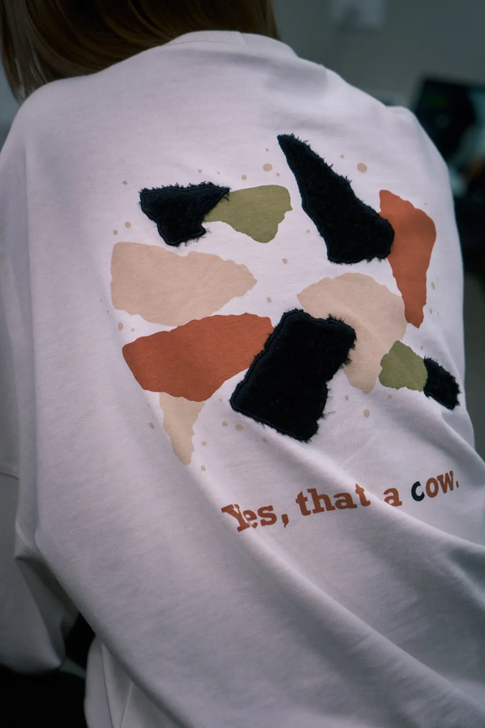 Close das costas de uma camiseta off-white: manchas em terracota, preto, areia e verde, e abaixo delas a frase “Yes, that’s a cow.”