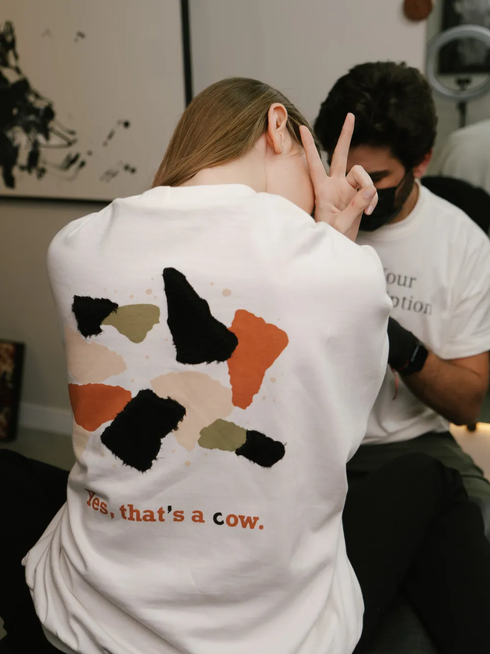 Duas pessoas num ateliê: uma de costas com a estampa da vaca, outra com a “Fuck Your Job Description”.