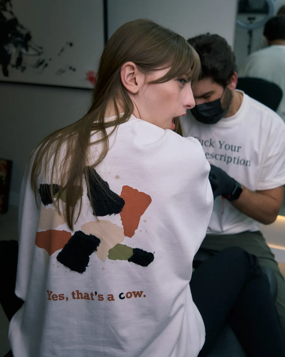 Duas pessoas de costas em um ateliê, uma com a camiseta da vaca e outra com a “Fuck Your Job Description”.